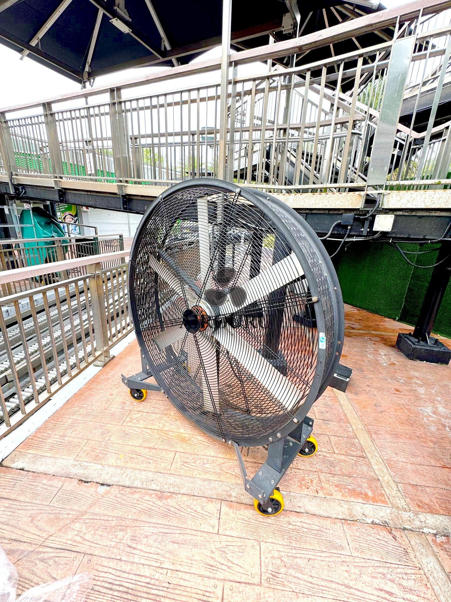 Airguru floor fan on a tiled attraction deck beside queue railings