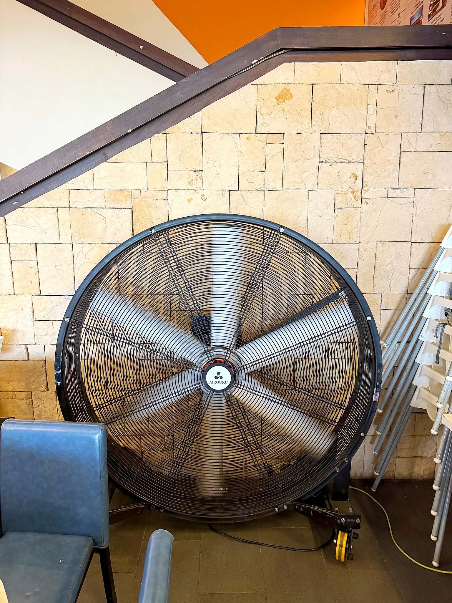 Large industrial floor fan beside a stone wall and restaurant chairs