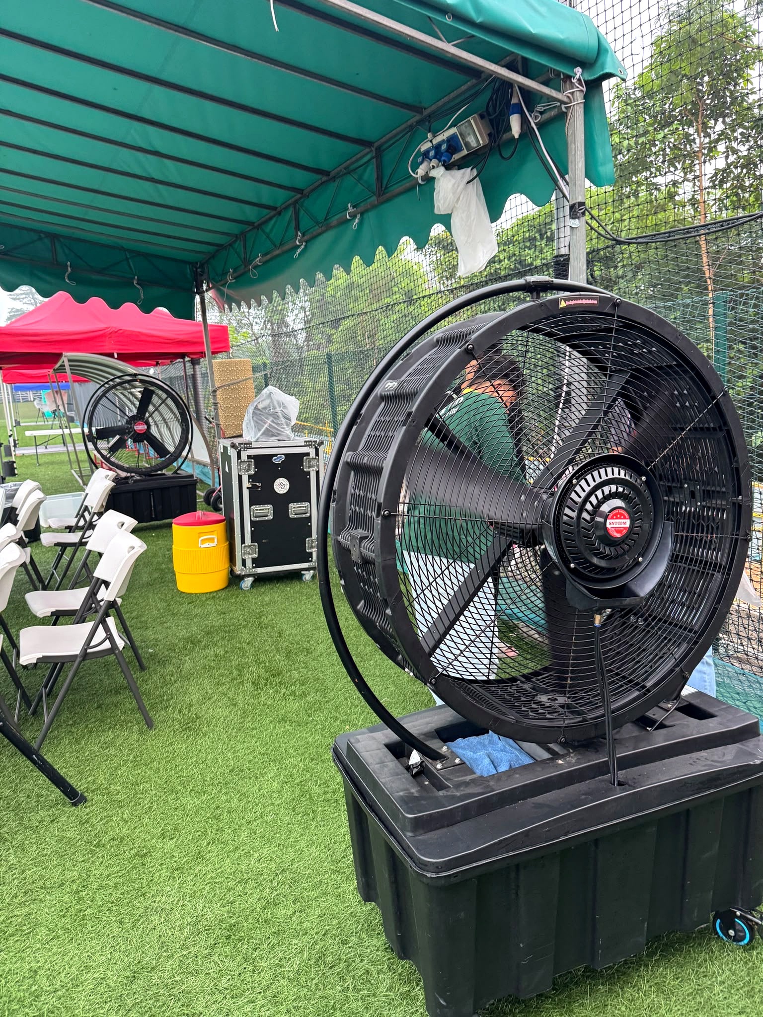 Portable fan on a wheeled base beside chairs under a green canopy